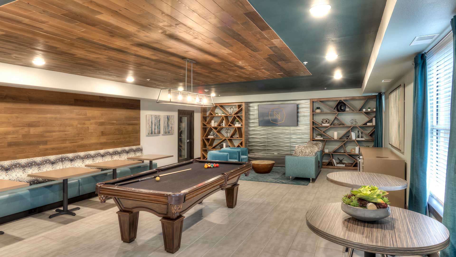 There is a pool table in the middle of the room. The far-left wall has bench seating with tables. The back wall is a seating area with couches. Café tables line the right wall near the camera.