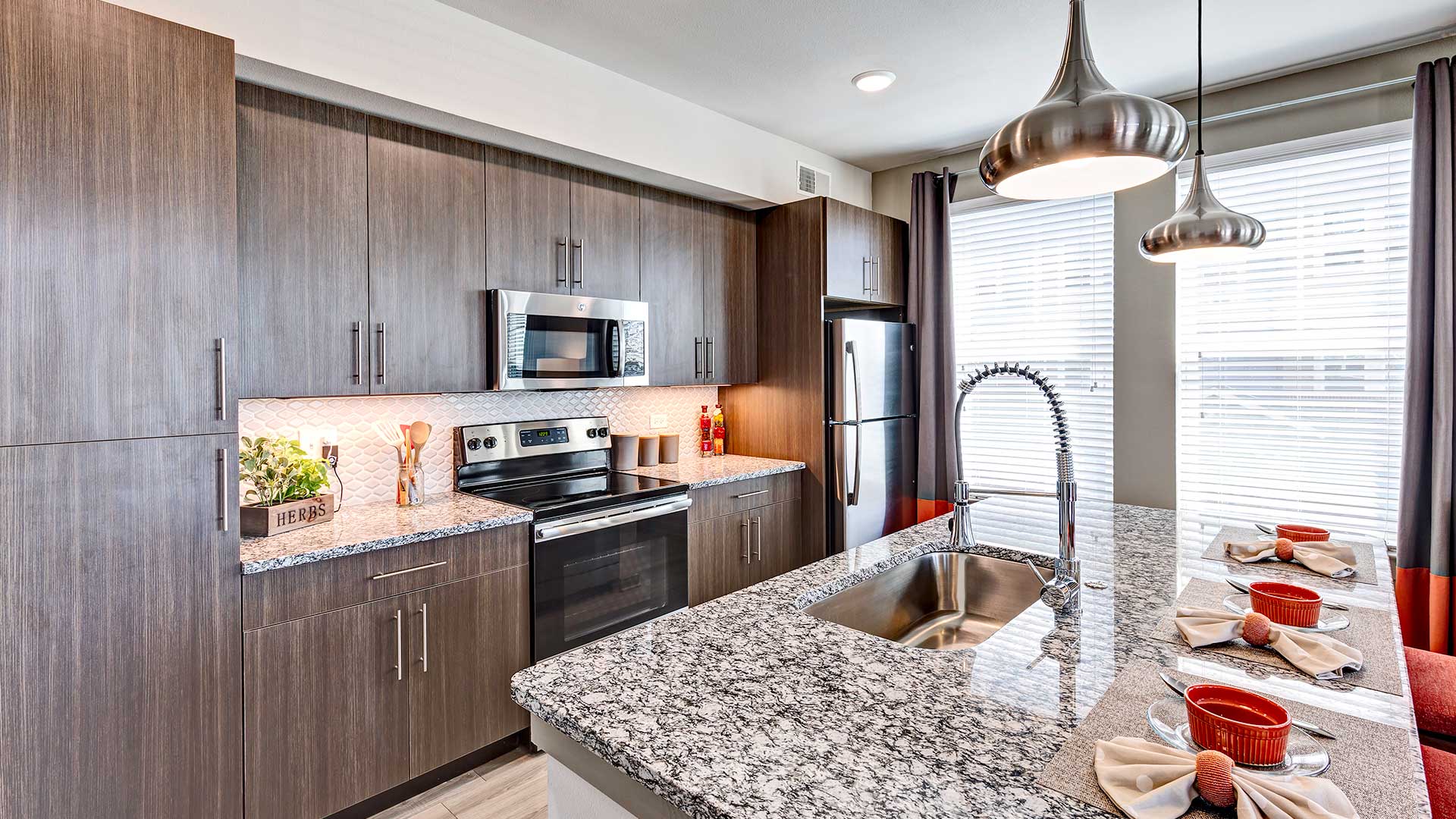 Looking at an angle across a kitchen island with three place settings along the right at the kitchen against the left wall. It has stainless steel appliances and brown cabinets.