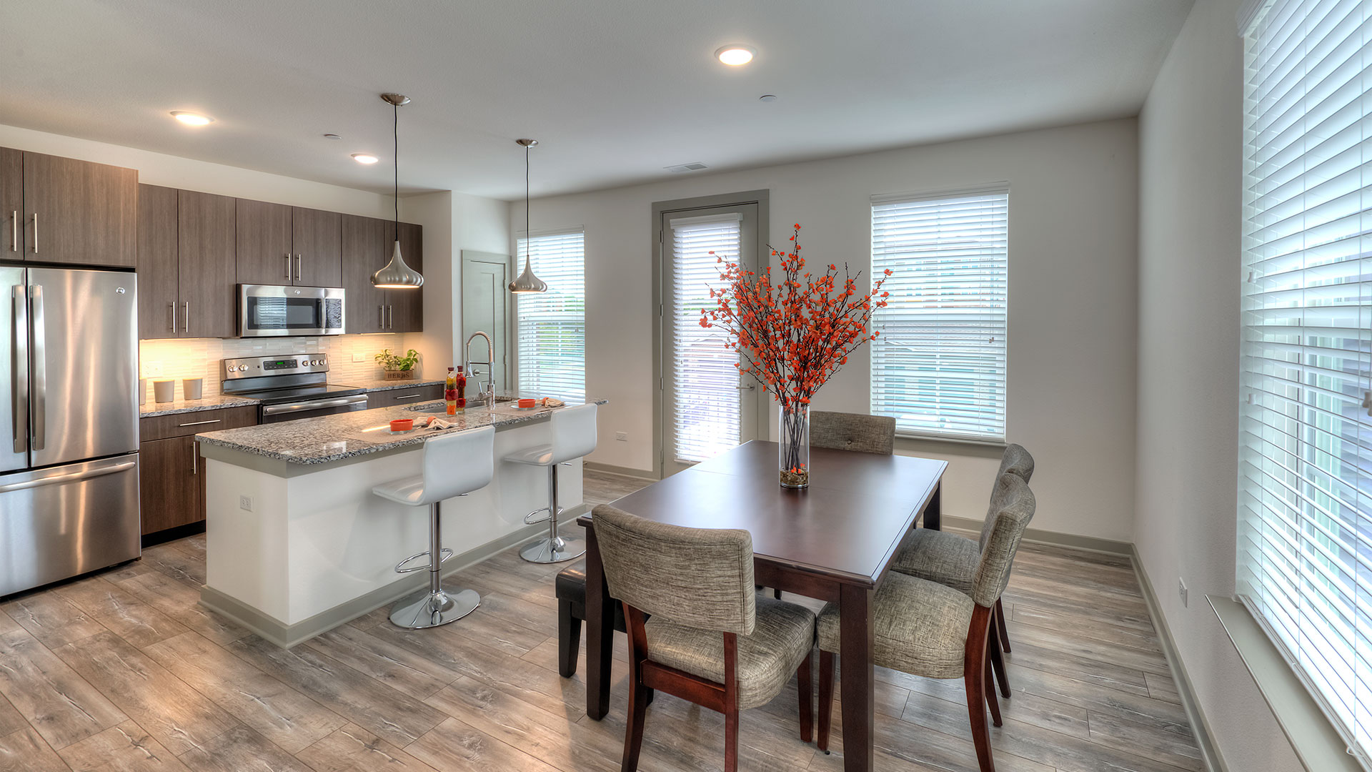 A kitchen with stainless steel appliances and brown cabinets lines the back left wall. An island is before it with two stools and place settings. On the right is a dining table with a large vase and flowers.